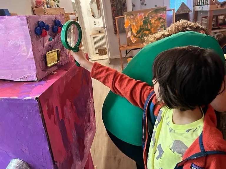 A child holds a magnifying glass up to the face of a robot built from cardboard boxes, painted purple and pink.