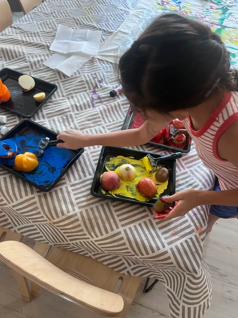 A child seen from above, pressing halved apples and onions into trays of blue and yellow paint.